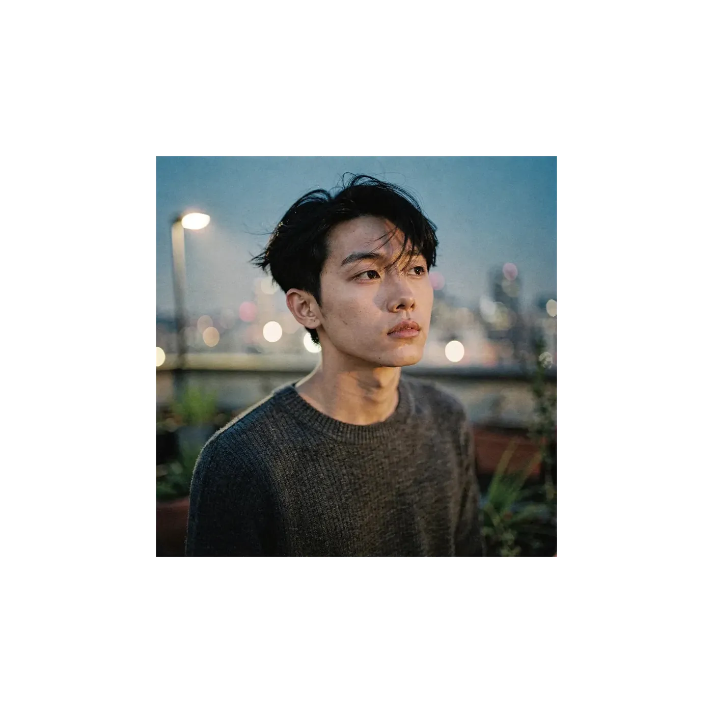 Square 1:1 photograph of a young man in a grey sweater on a city rooftop at dusk with bokeh lights behind him, the before frame of a vertical reframe, generated on getimg.ai.