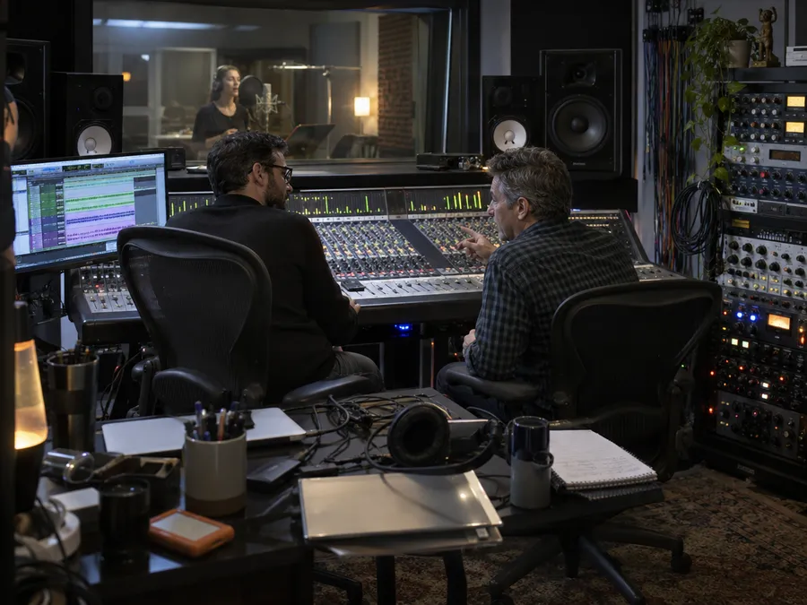 Two engineers at a large mixing console in a studio control room while a voice artist records in the booth behind the glass, showing traditional voice recording. Image generated on getimg.ai.