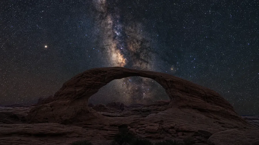 AI-generated star image: Milky Way galactic core rising over a red-rock desert arch at new moon.