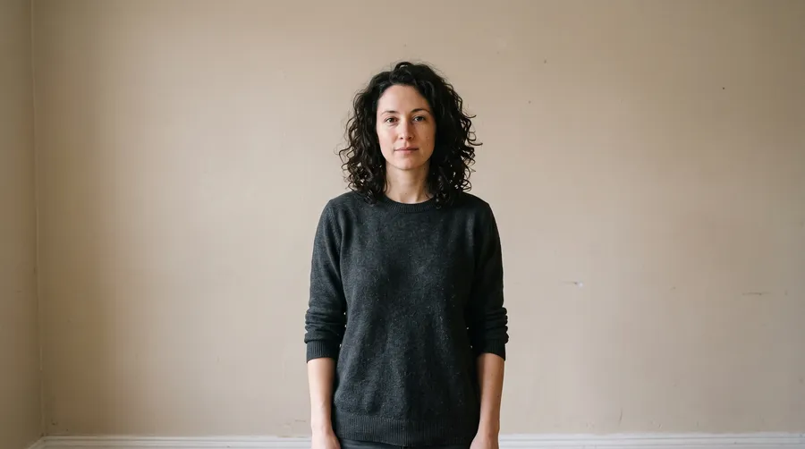 Portrait of a woman with dark curly hair against a plain beige wall, the source photo before an AI background change on getimg.ai
