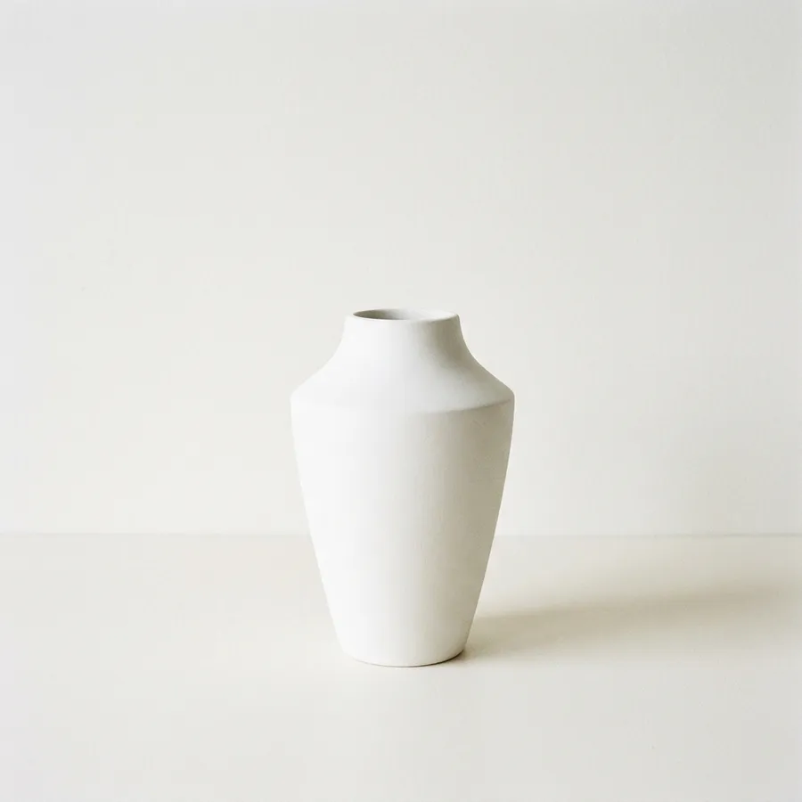 White matte ceramic vase standing alone on a pale off-white table against an off-white wall with almost no tonal separation, the original image before background removal, generated with getimg.ai