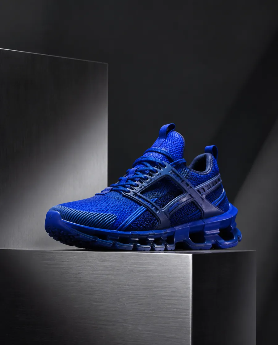 a cobalt-blue technical sneaker, placed on a muted brushed-steel platform, dark graphite surroundings, controlled soft spotlight, realistic material details and clear product hierarchy, Pinterest Gen Z editorial aesthetic, horizontal 16:9, realistic product photo generated on getimg.ai