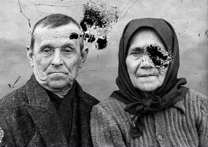 A black-and-white portrait of an elderly man and a woman in a headscarf, marked by dark ink blots across his forehead and her cheek: the damaged original before AI restoration on getimg.ai.