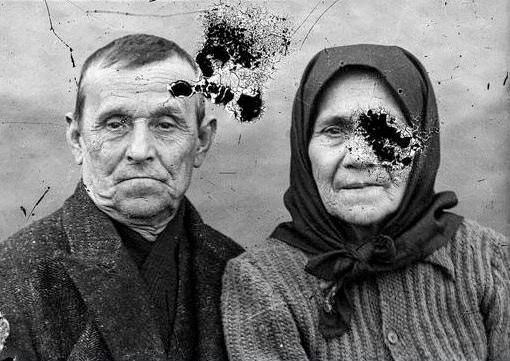A black-and-white portrait of an elderly man and a woman in a headscarf, marked by dark ink blots across his forehead and her cheek: the damaged original before AI restoration on getimg.ai.