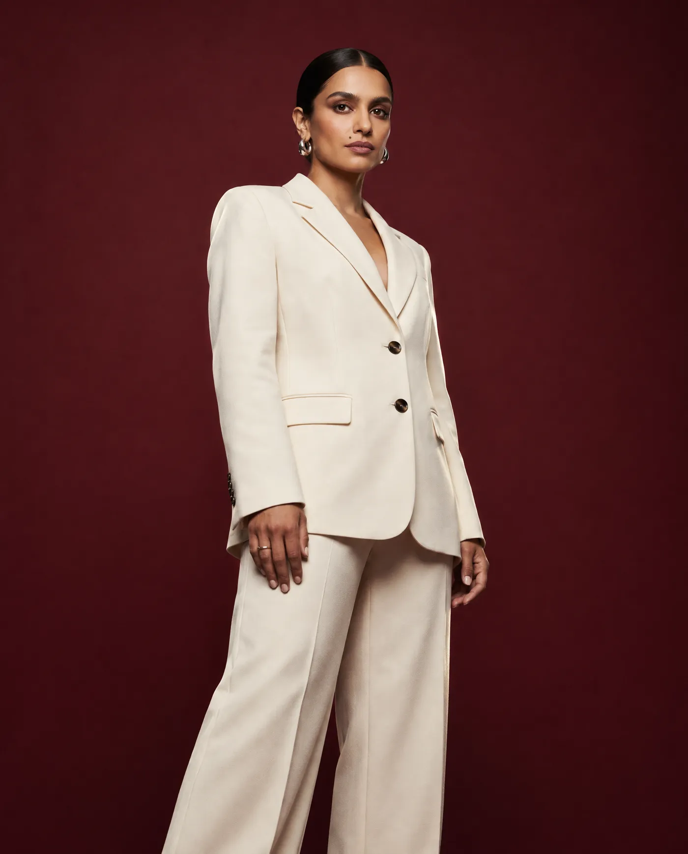 Shot 8, low-angle portrait from below eye level: a fictional model with a sleek low bun and silver hoop earrings in a cream tailored suit on a burgundy studio backdrop, hand resting at her side, generated on getimg.ai in a guide to creating an AI photoshoot from one photo