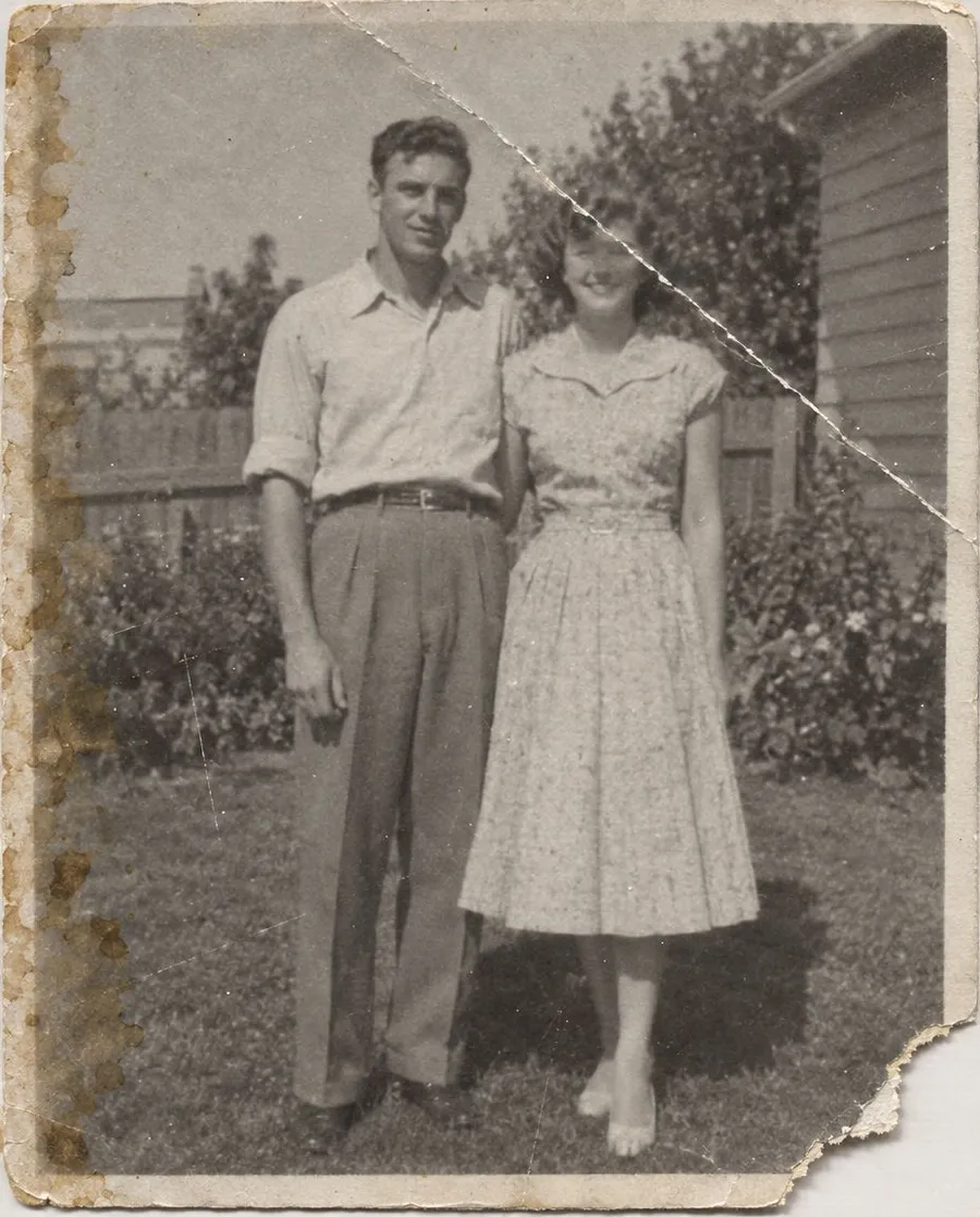 Before AI photo restoration: a creased, faded 1950s black-and-white snapshot of a young couple standing on a lawn, with water stains and a torn edge down the left side and a diagonal crack across the print. Example damaged input for the Photo restoration Action on getimg.ai.