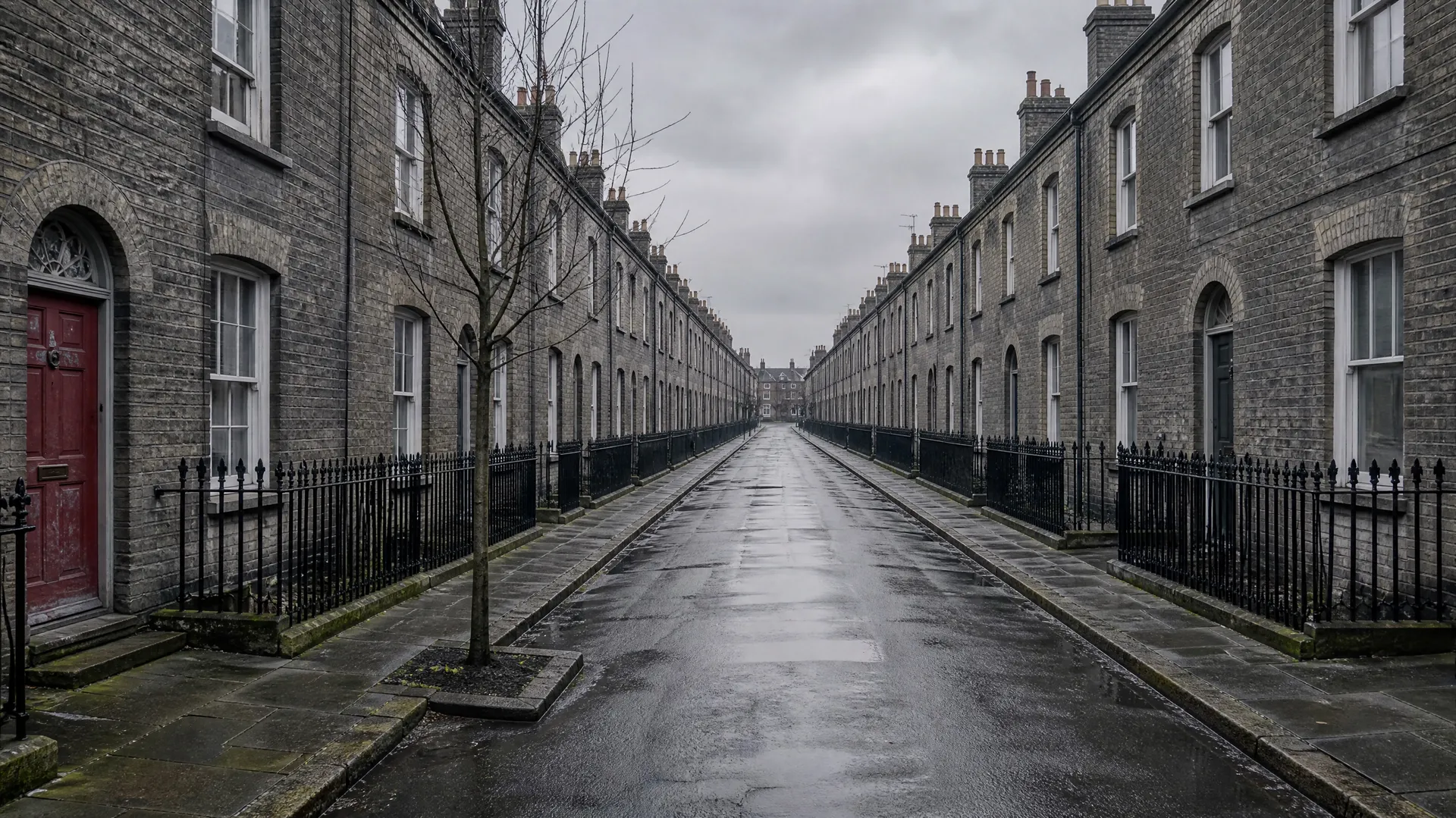 Empty residential street lined with grey brick terraced houses and black railings on a wet, overcast day.