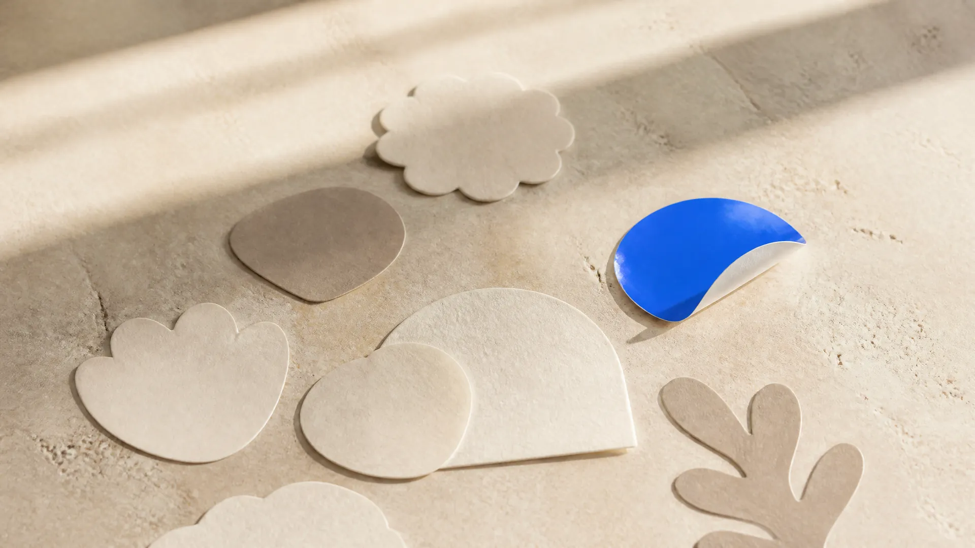 Matte die-cut sticker shapes in cream tones on a warm surface with one glossy electric-blue sticker peeling up, representing the AI sticker maker. Generated with getimg.ai.