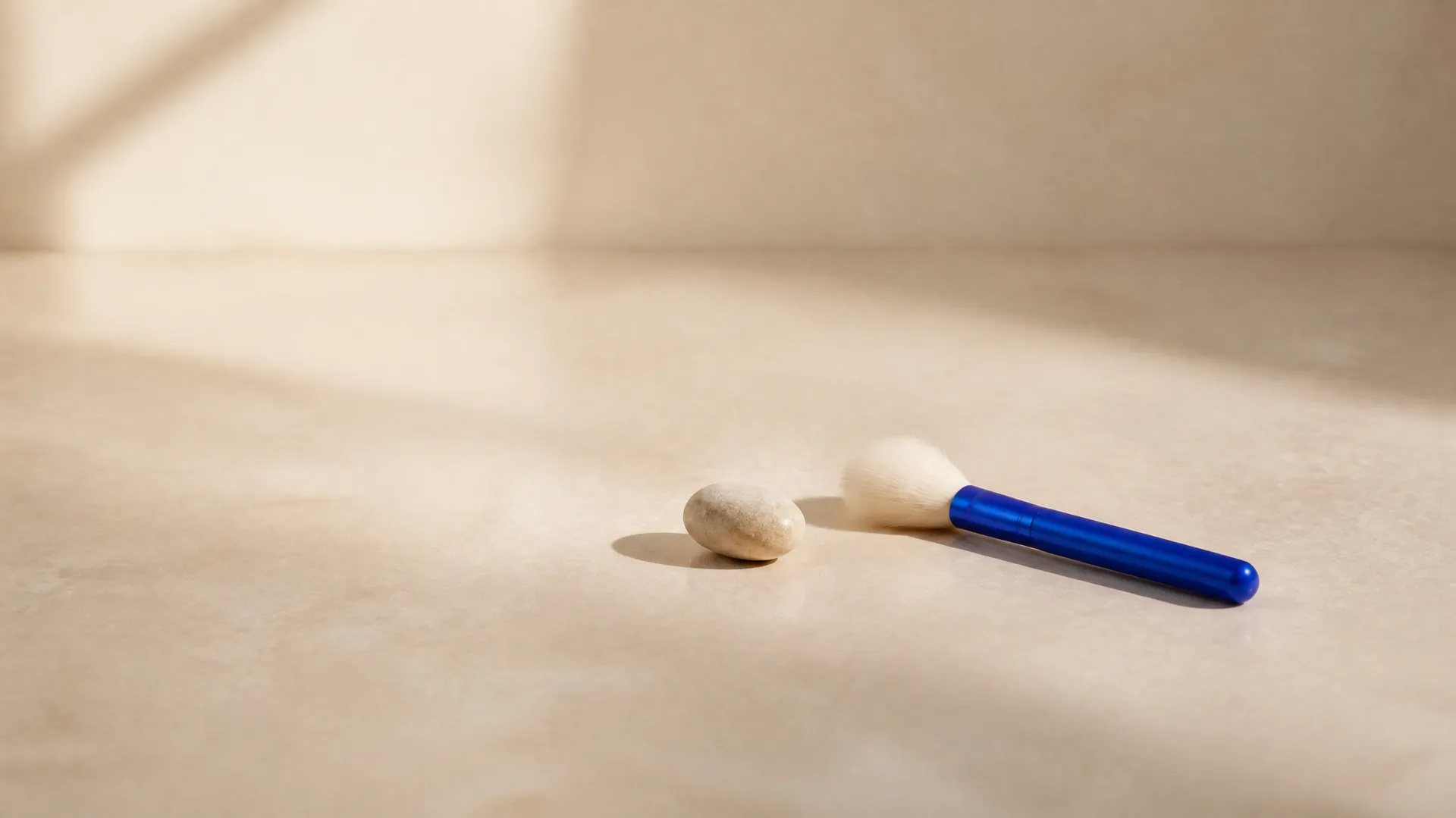 A clean cream tabletop bare except for a smooth stone and a dust brush with an electric-blue handle, representing AI removal of unwanted objects from photos. Generated with getimg.ai.