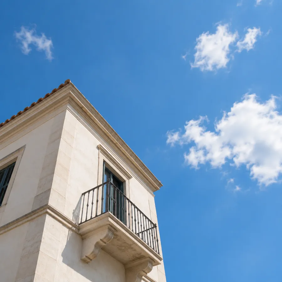 Corner of a pale stucco building with a small balcony seen from below against a blue sky with white clouds.