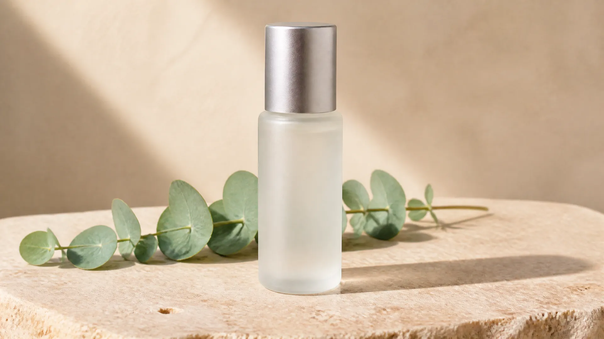 Frosted bottle with a silver cap beside eucalyptus leaves on a sunlit stone slab.