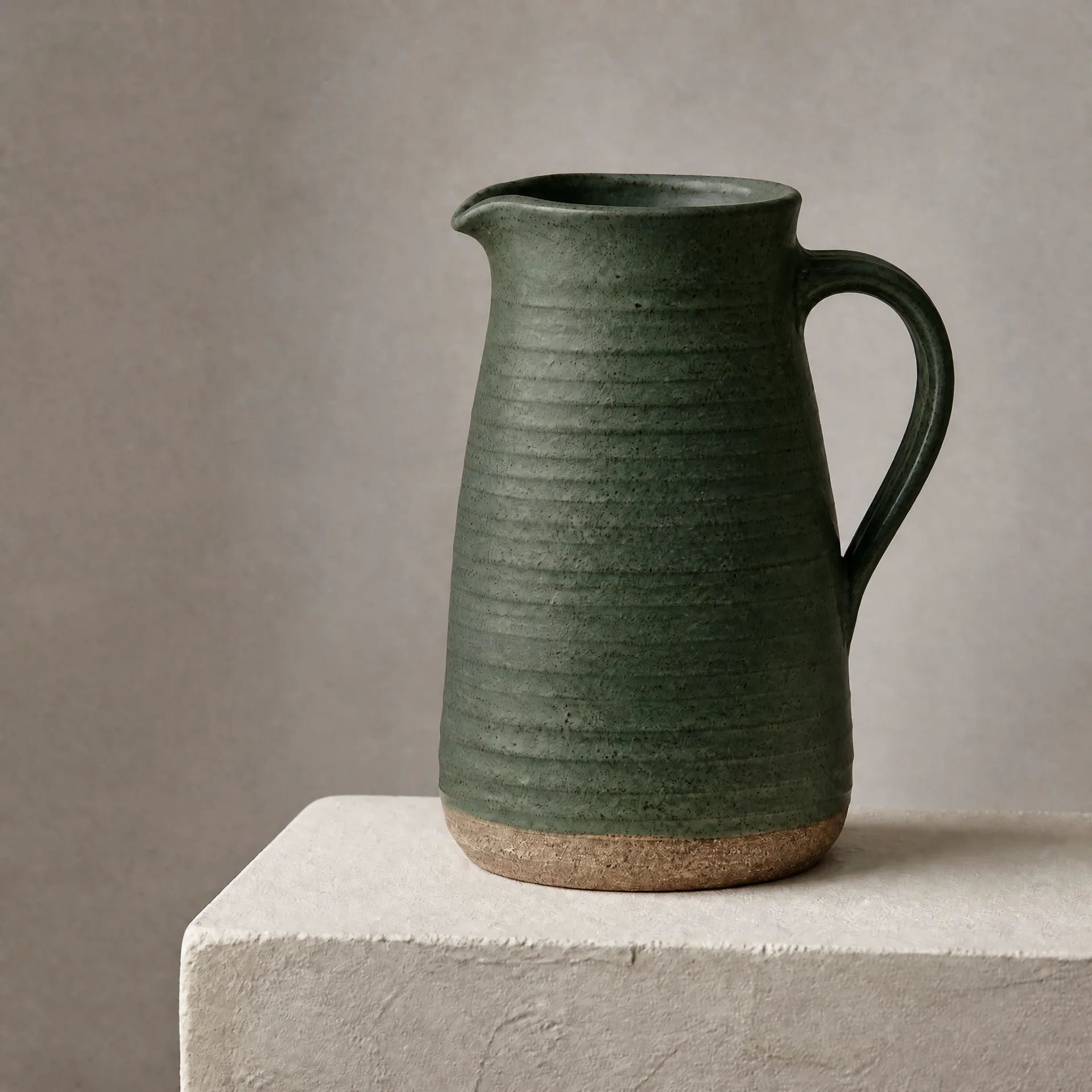 Dark green ceramic jug with a curved handle and unglazed base on a pale square plinth.