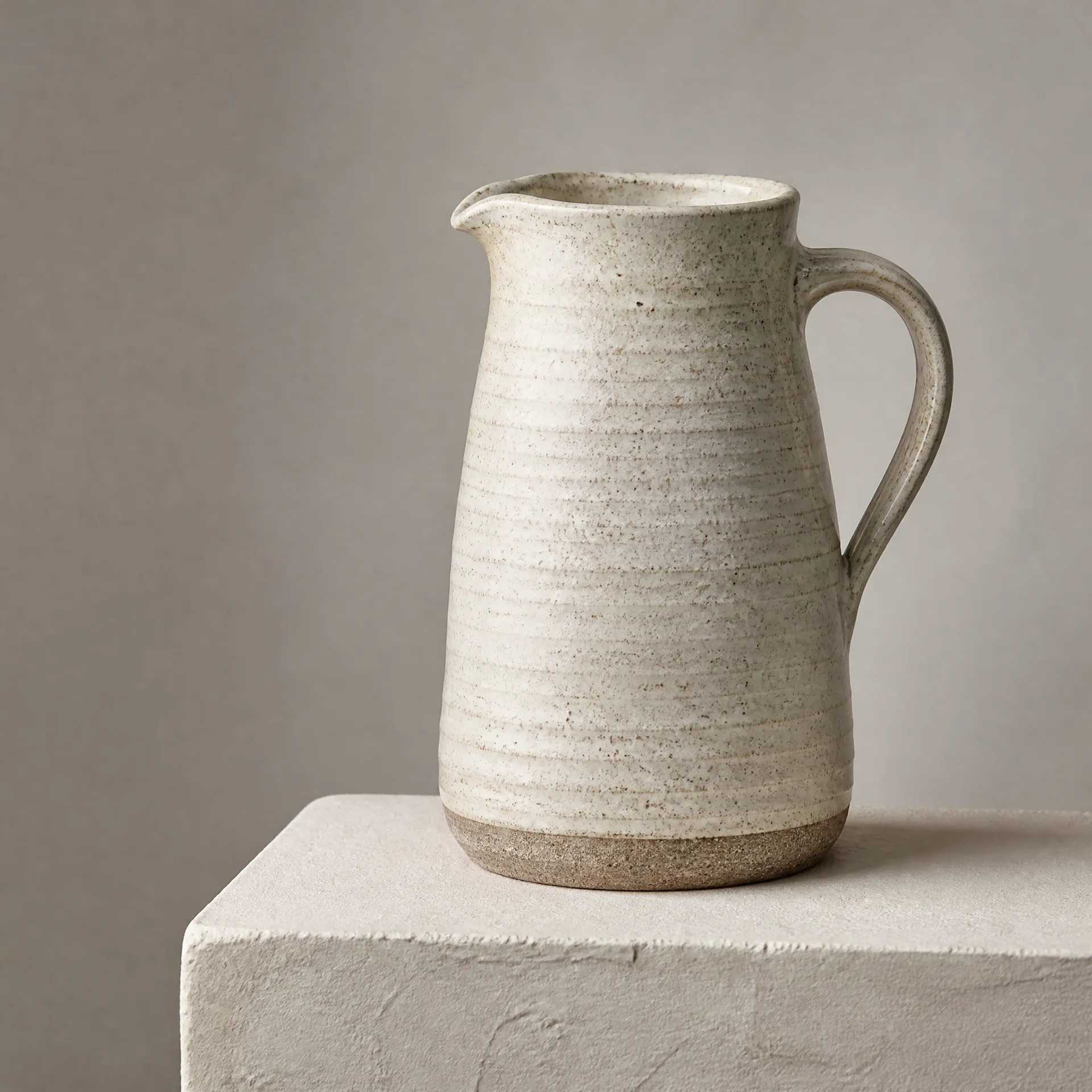 Speckled cream ceramic jug with a curved handle on a pale square plinth.