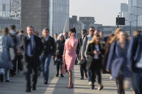 Before AI object removal: a woman in a pink dress talks on her phone while standing still on a city bridge, surrounded by a blurred crowd of business commuters walking past a modern skyline. Example photo used with getimg.ai to show a busy background that needs distractions removed.