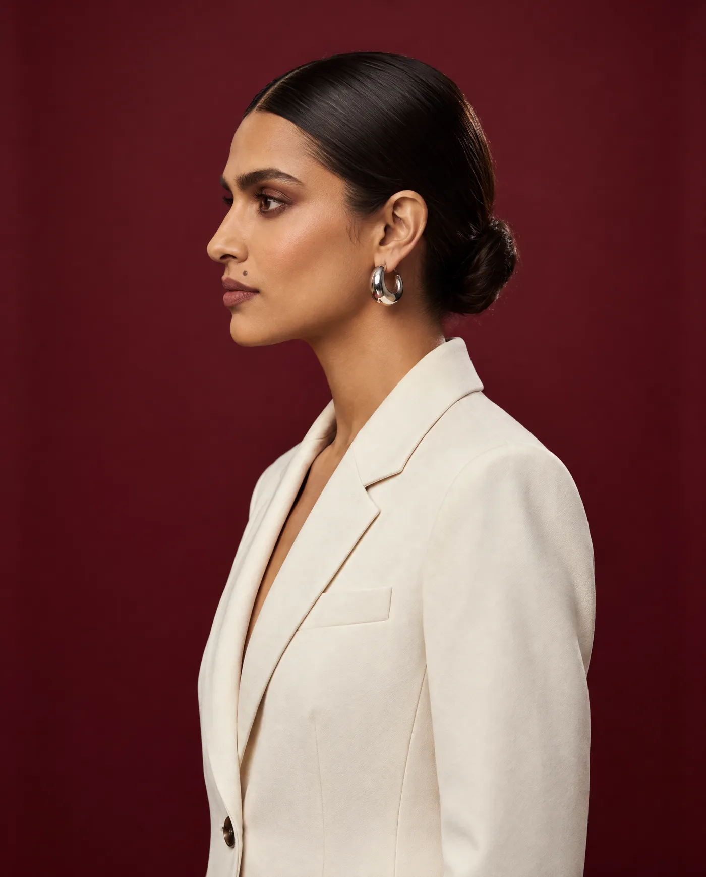 Shot 5, clean left profile on 85mm: a fictional model with a sleek low bun and silver hoop earrings in a cream tailored suit on a burgundy studio backdrop, showing the bun and earring from the side, generated on getimg.ai in a guide to creating an AI photoshoot from one photo