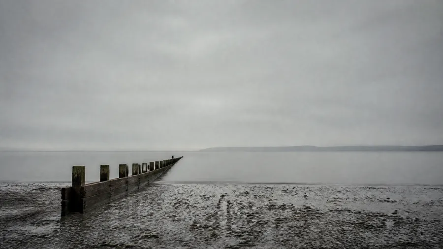 Empty shingle beach with a long wooden groyne extending into calm grey water beneath an overcast, misty sky.