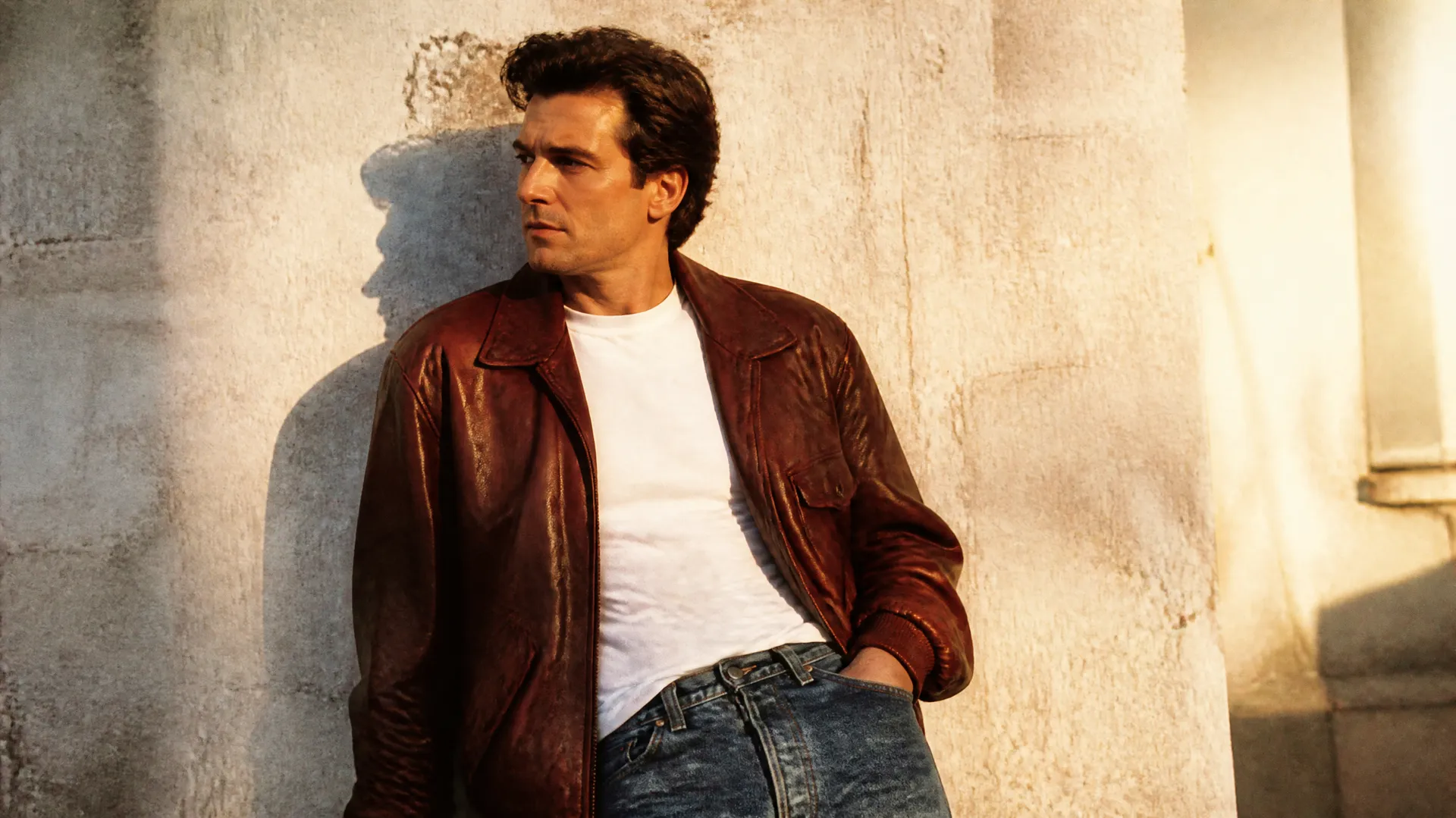 Man in a brown leather jacket, white T-shirt and jeans leaning against a sunlit textured wall.