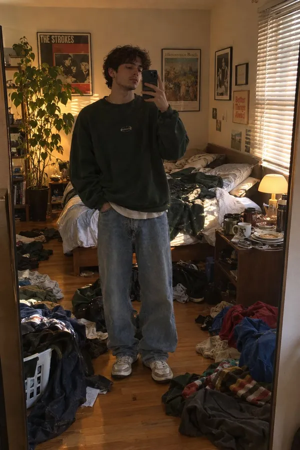 Before AI object removal: a young man in a green sweatshirt takes a phone mirror selfie in a warmly lit bedroom cluttered with piles of clothes across the wooden floor, a laundry basket, an unmade bed, and a nightstand covered in mugs and dishes. Example photo made with getimg.ai showing clutter that needs cleaning up.