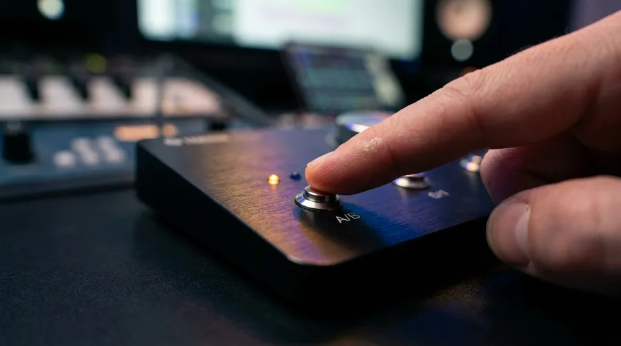 Close-up of a finger pressing an A/B comparison button on a studio monitor controller with amber and blue indicator lights, representing switching between two music models. Image generated with getimg.ai.
