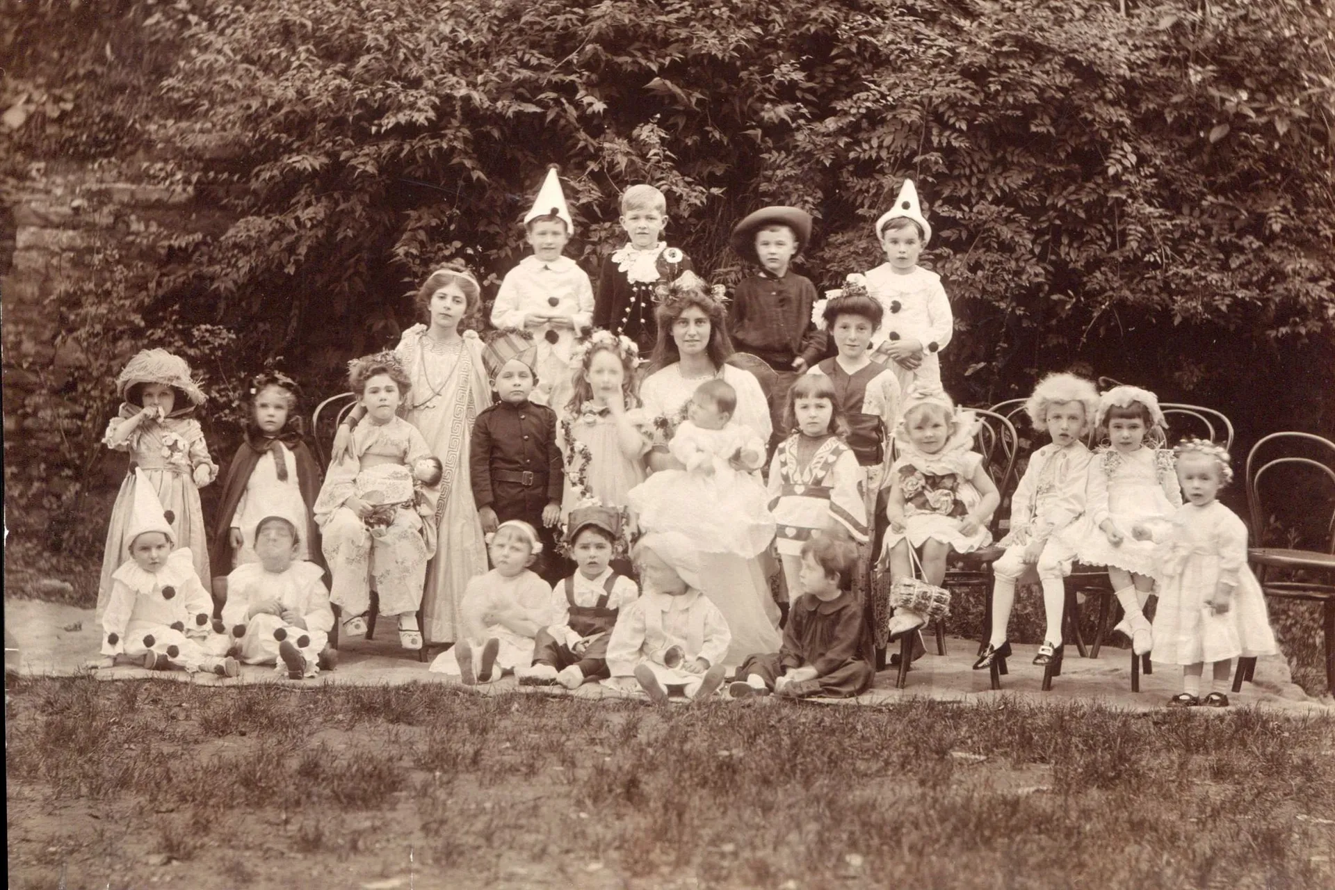 A faded sepia group photograph of many children in vintage costumes and pointed party hats posing outdoors: the aged original before AI restoration on getimg.ai.