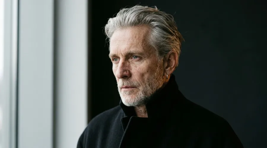 Editorial portrait of a silver-haired man around 64 in a black high-collar coat, lit by soft window light against a charcoal background, generated on getimg.ai with Nano Banana 2 for an AI image realism test.