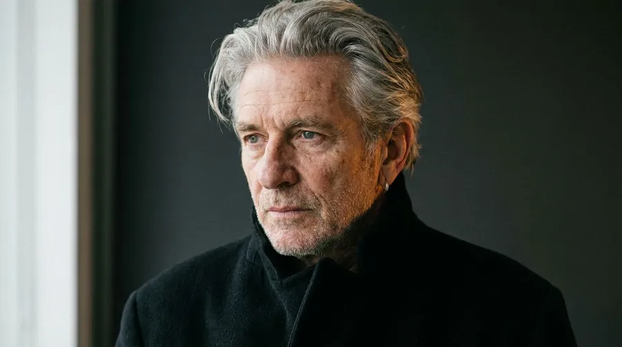 Editorial portrait of a silver-haired man around 64 in a black high-collar coat, lit by soft window light against a charcoal background, generated on getimg.ai with Nano Banana Pro for an AI image realism test.