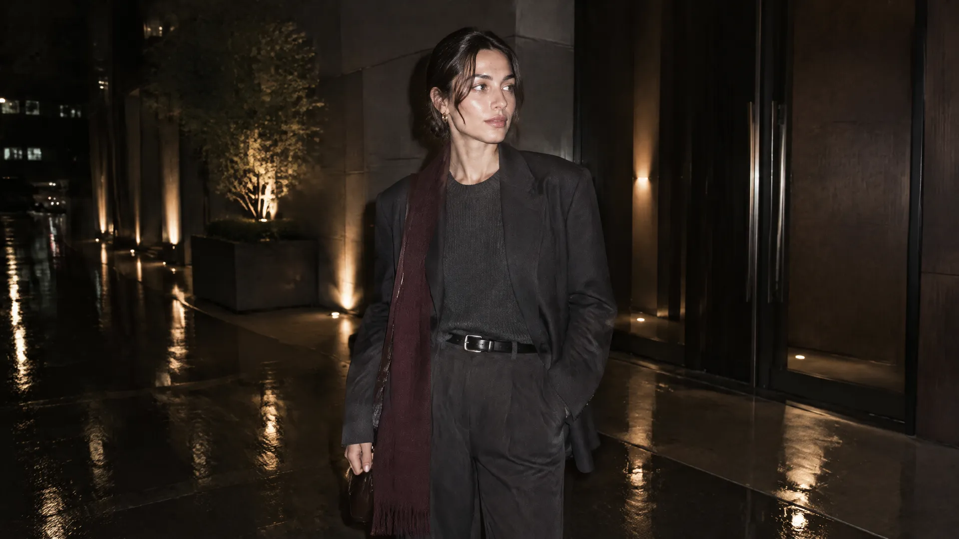 An AI-generated night shot of Mara in a dark graphite ensemble and burgundy scarf on a rain-slicked street, the Round 5 low-light test that keeps her palette intact; created on getimg.ai.