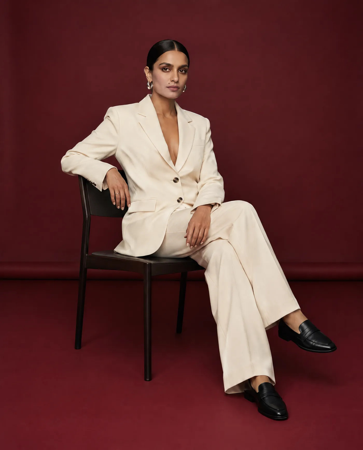 Shot 9, seated pose: a fictional model with a sleek low bun and silver hoop earrings in a cream tailored suit on a burgundy studio backdrop sitting on a minimal black chair, generated on getimg.ai in a guide to creating an AI photoshoot from one photo