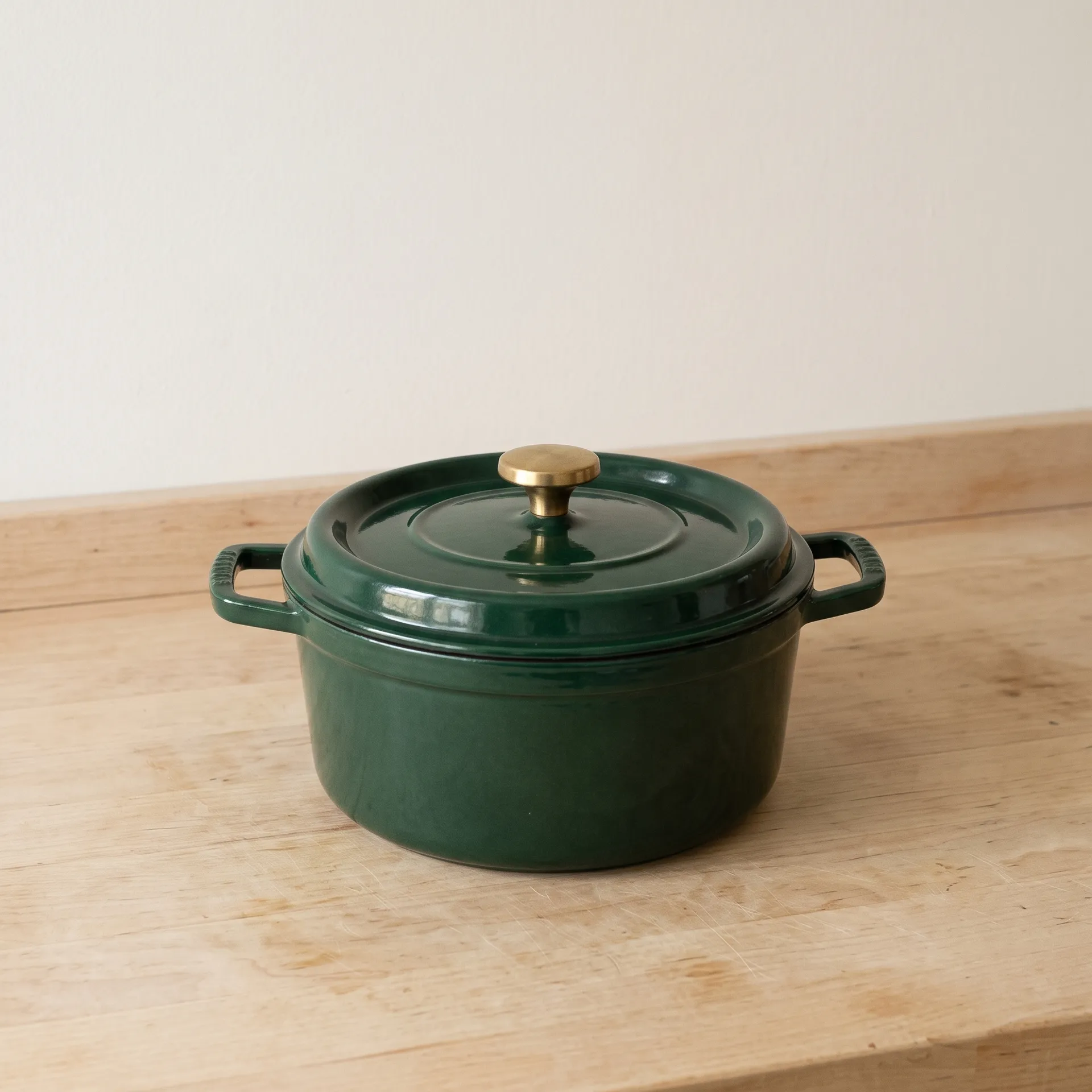 A deep green enamelled cast iron casserole pot with a brass knob standing on a pale wooden kitchen counter in soft daylight, plain wall behind, no props. An ordinary product photo that is not yet listing ready, generated in getimg.ai.