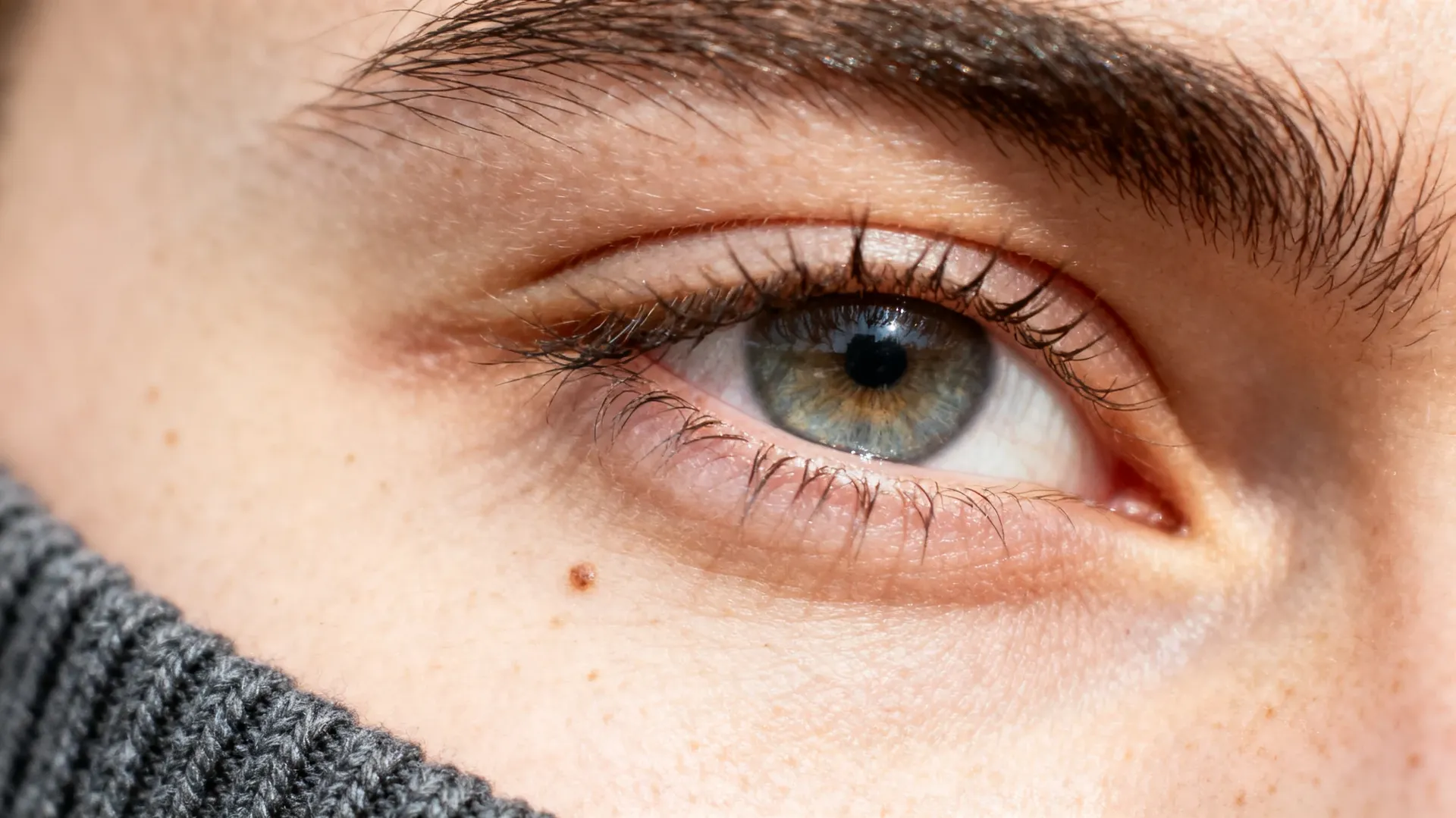 Extreme macro of a human eye showing individual lashes, eyebrow hairs, skin pores, a small mole and a softbox catchlight in the iris, with a knitted collar at the edge of frame. The level of detail every AI edit was reviewed at before delivery, generated with getimg.ai.