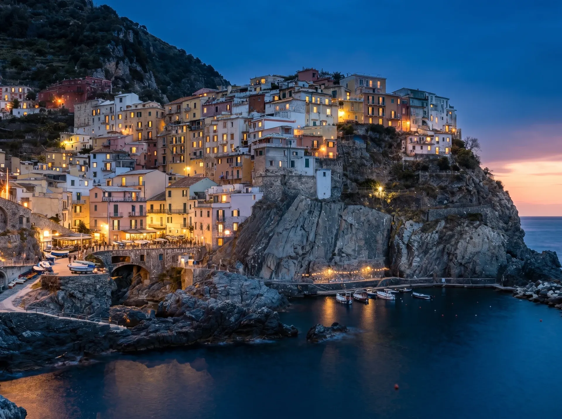 The same hillside town at blue hour, with warm window lights and harbour lamps switched on under a deep blue sky. Time-of-day edit made with Nano Banana 2 on getimg.ai.