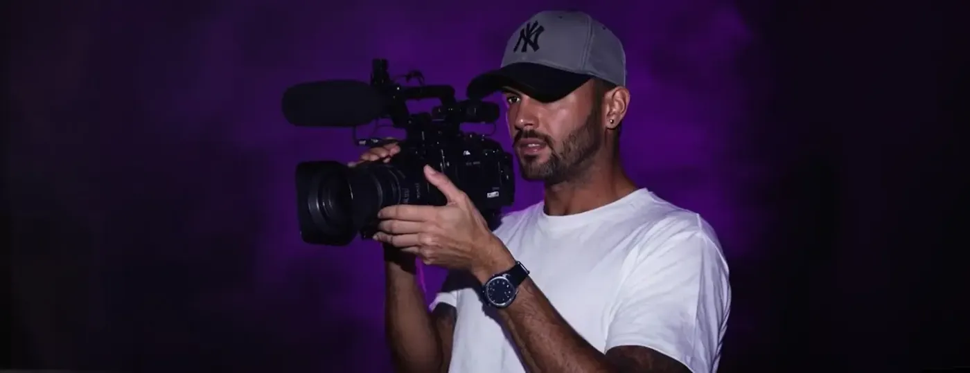 A man in a baseball cap and white T-shirt holds a video camera against a purple-lit background.