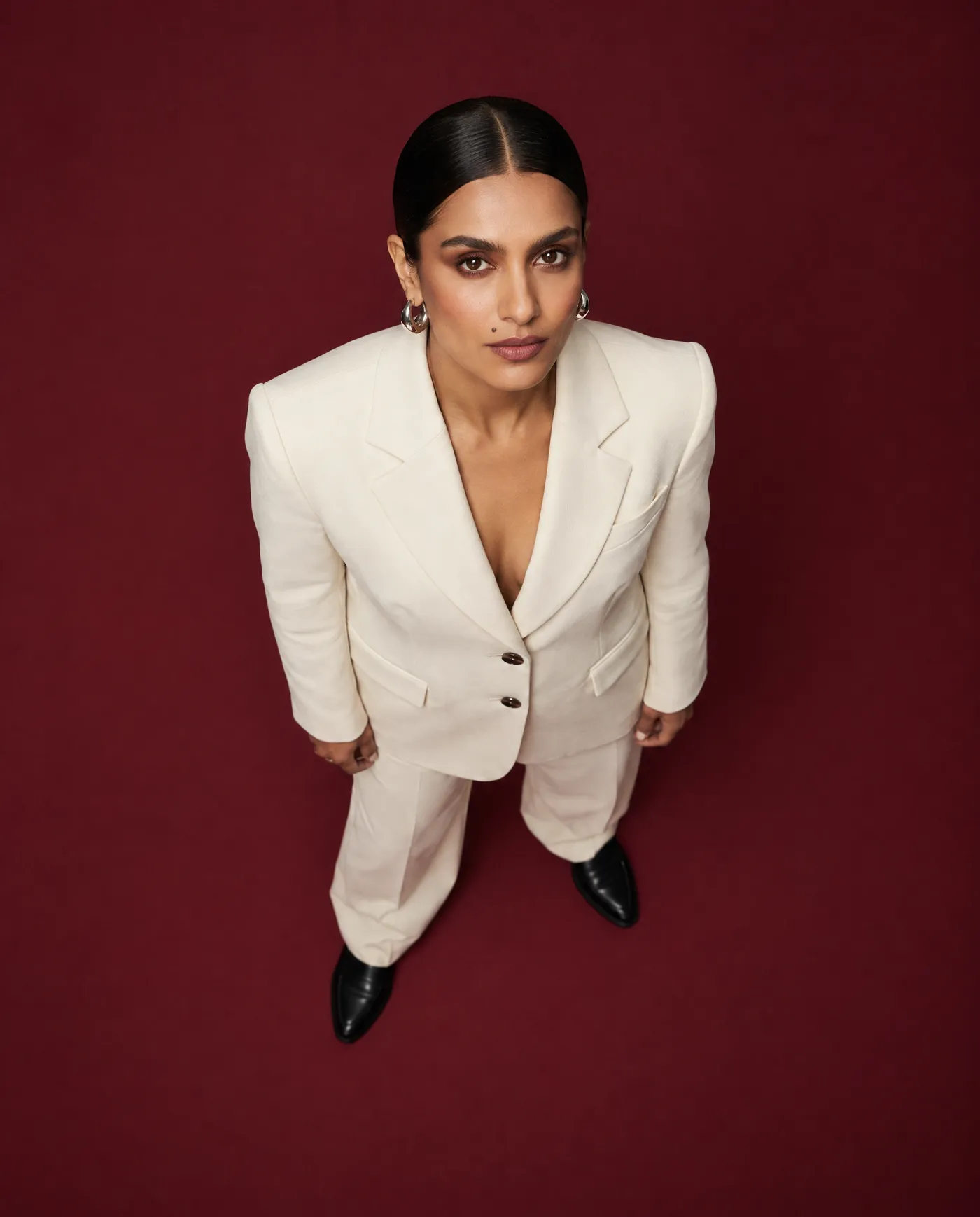 Shot 7, true overhead top-down view: a fictional model with a sleek low bun and silver hoop earrings in a cream tailored suit on a burgundy studio backdrop, looking up at the camera from the burgundy floor, generated on getimg.ai in a guide to creating an AI photoshoot from one photo