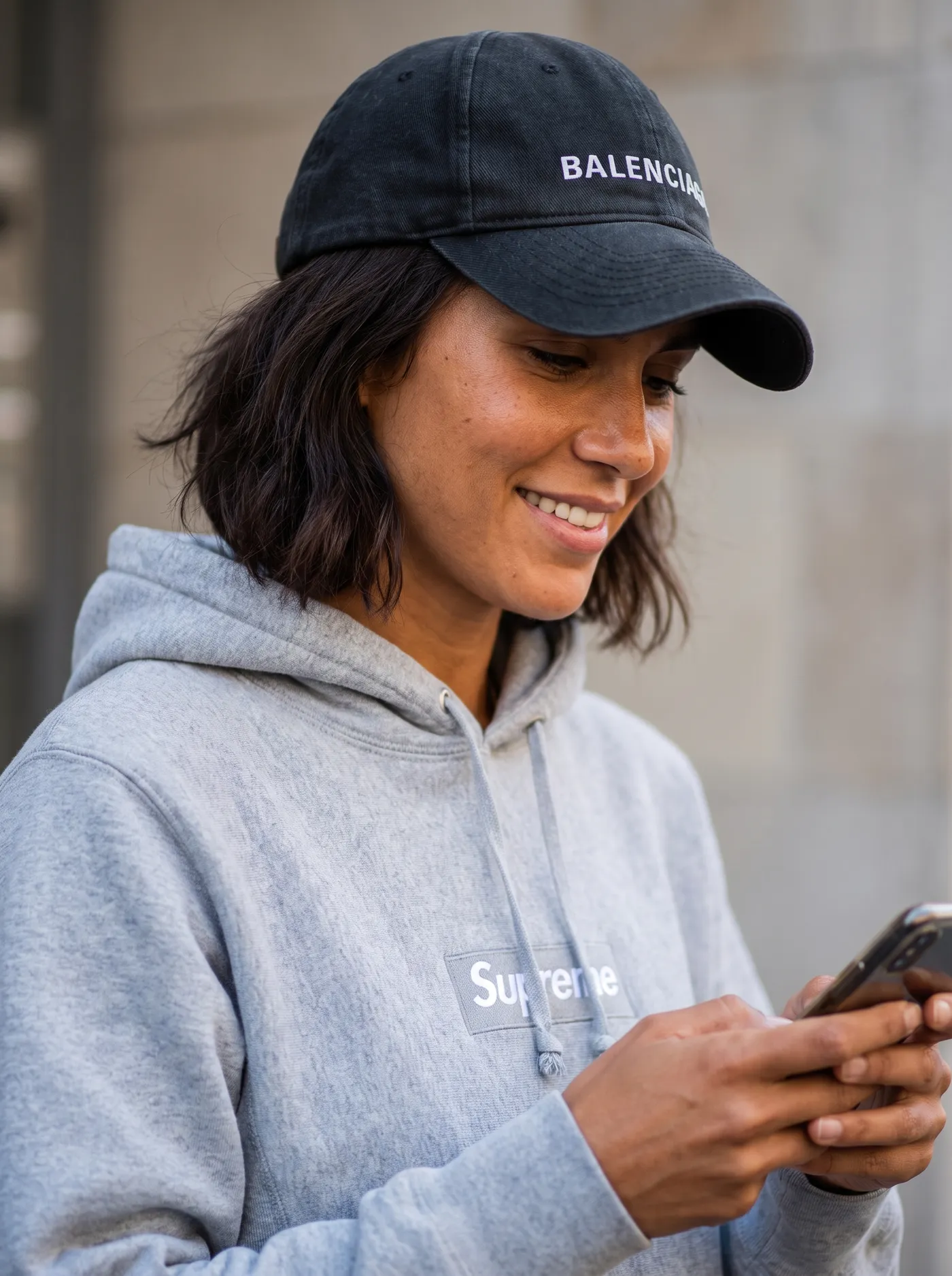 Character creator result on getimg.ai: a photorealistic young woman with a dark bob smiling down at her phone, in a black logo cap and grey hoodie on a blurred city street. Generated from chosen attributes rather than an uploaded photo.
