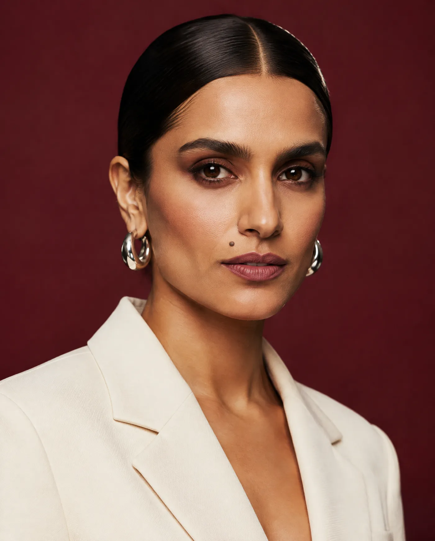 Shot 11, tight facial close-up on 85mm with sharp eyes and real skin texture: a fictional model with a sleek low bun and silver hoop earrings in a cream tailored suit on a burgundy studio backdrop, generated on getimg.ai in a guide to creating an AI photoshoot from one photo