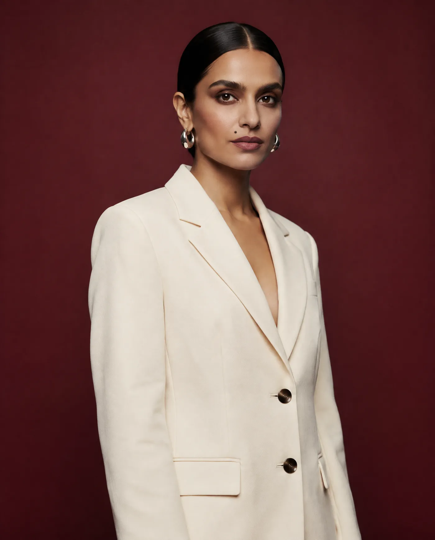 Shot 3, three-quarter waist-up on 50mm: a fictional model with a sleek low bun and silver hoop earrings in a cream tailored suit on a burgundy studio backdrop, torso turned slightly, generated on getimg.ai in a guide to creating an AI photoshoot from one photo