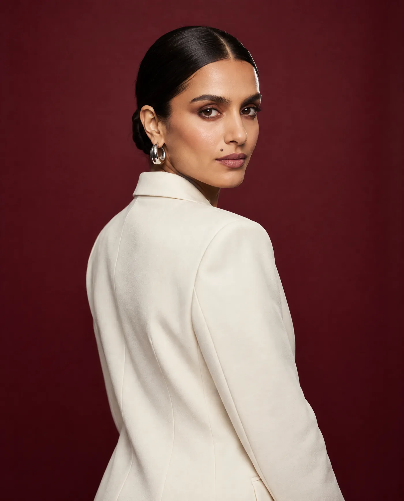 Shot 6, over-the-shoulder: a fictional model with a sleek low bun and silver hoop earrings in a cream tailored suit on a burgundy studio backdrop, body turned away and head looking back at the camera, generated on getimg.ai in a guide to creating an AI photoshoot from one photo