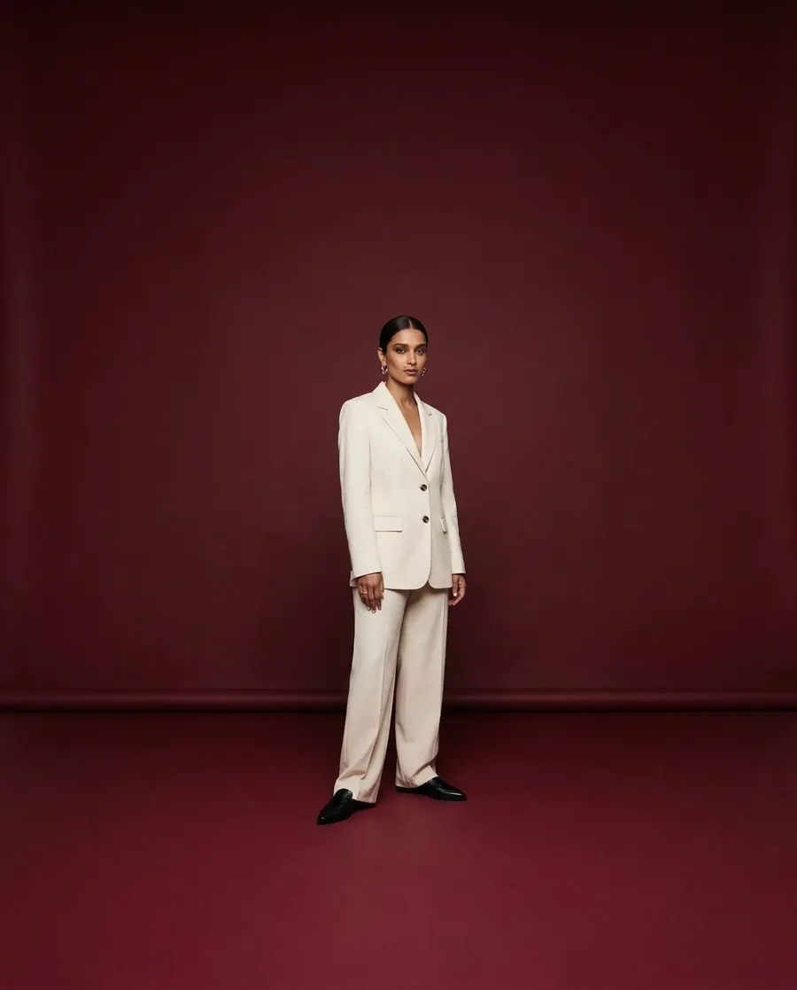 Shot 1, wide establishing shot on a 35mm lens: a fictional model with a sleek low bun and silver hoop earrings in a cream tailored suit on a burgundy studio backdrop, standing small in frame with the seamless backdrop and floor visible, generated on getimg.ai in a guide to creating an AI photoshoot from one photo