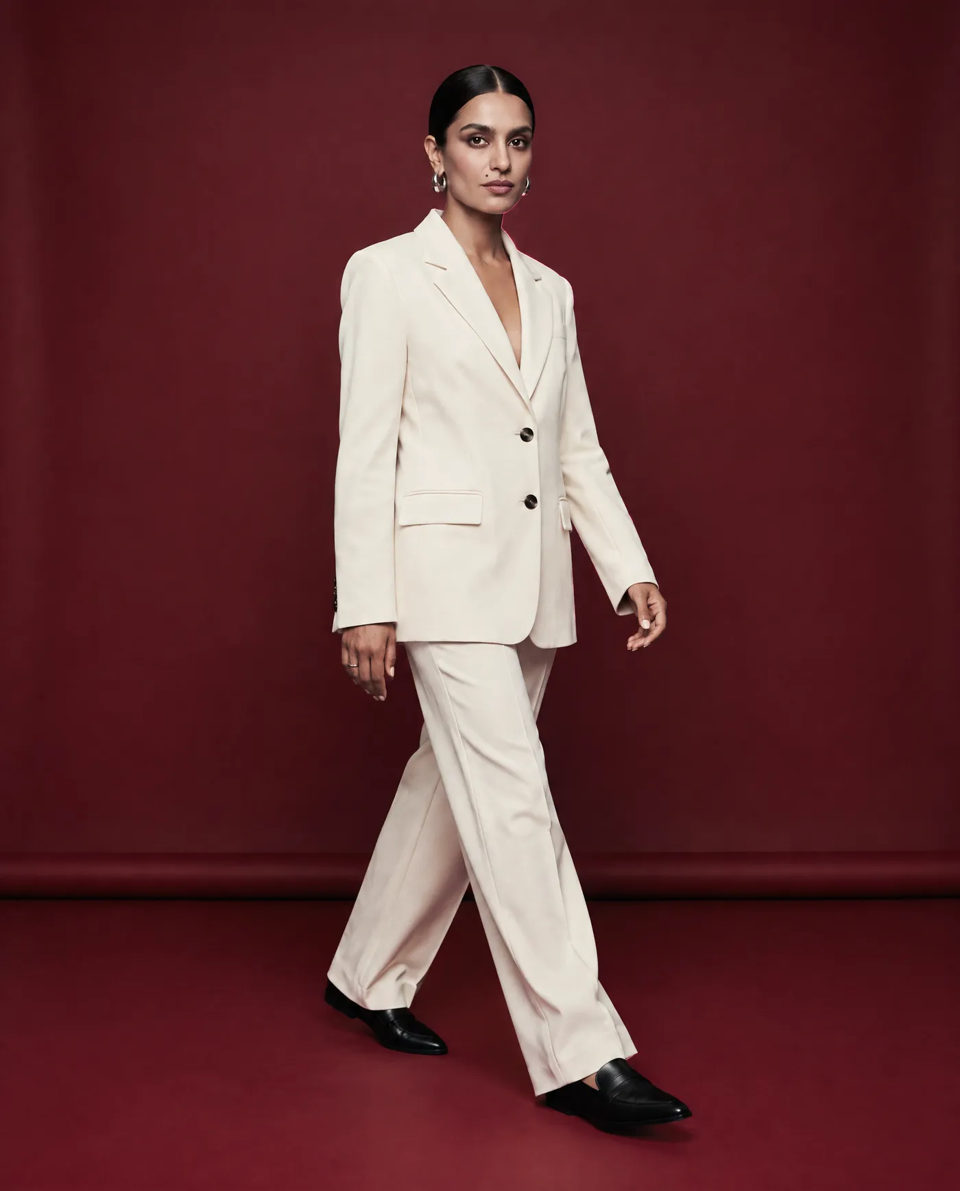 Shot 10, full-body walking shot mid-stride: a fictional model with a sleek low bun and silver hoop earrings in a cream tailored suit on a burgundy studio backdrop and black loafers, generated on getimg.ai in a guide to creating an AI photoshoot from one photo
