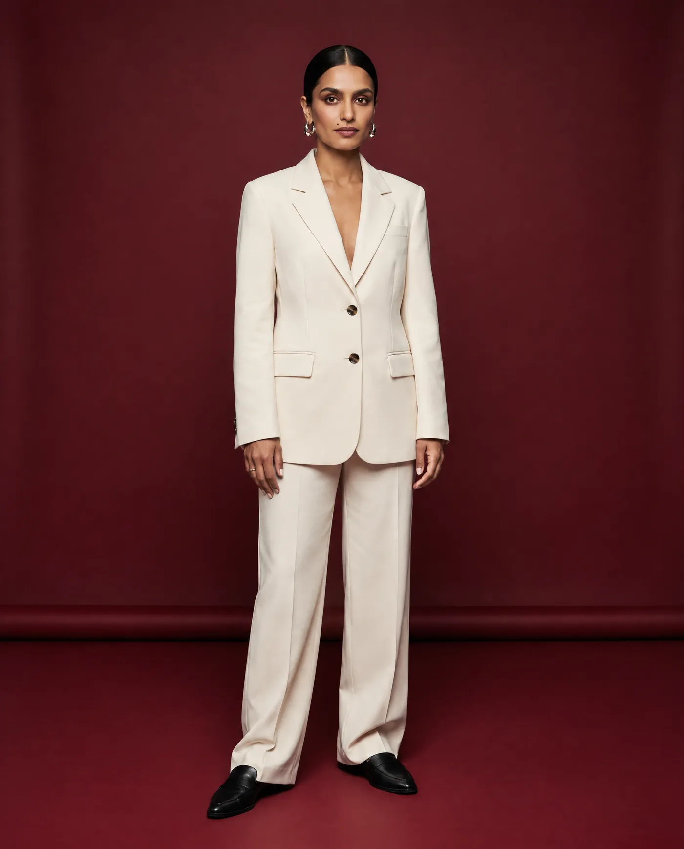 Shot 2, full-body front at eye level on 50mm: a fictional model with a sleek low bun and silver hoop earrings in a cream tailored suit on a burgundy studio backdrop and black loafers, the wardrobe check, generated on getimg.ai in a guide to creating an AI photoshoot from one photo